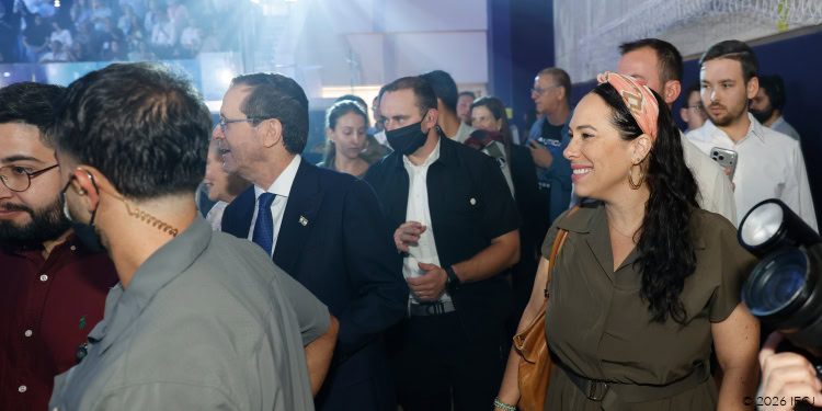 Group of people at a public event with media, cameras, and security; a smiling woman in a brown dress stands among attendees.