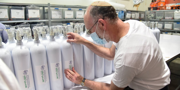Man wearing a kippah and facemask applies stickers showing the IFCJ logo onto white canisters of oxygen.
