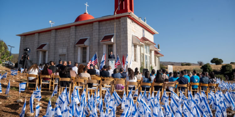 Gathering of people at a memorial service outside a church, with American and Israeli flags displayed prominently.