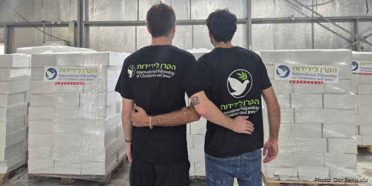 Two young men with arms around each other in a warehouse, wearing black T-shirts with the IFCJ logo, symbolizing support and unity for Christian aid efforts.
