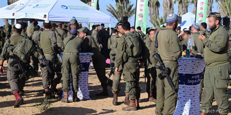Group of Israeli soldiers in uniform gathered around tables at an outdoor event, some holding rifles, with tents and palm trees in the background.