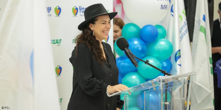Yael Eckstein speaking. black dress, black hat, microphone, podium, smiling, happy