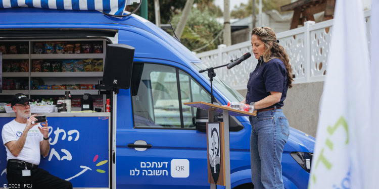 Fellowship Soldier Appreciation Trucks - Launching Ceremony - The Association for Israel’s Soldiers - Israel - April 2, 2025 woman in dark blue shirt speaking at podium, blue hospitality van in rear - man with beard sitting with camera