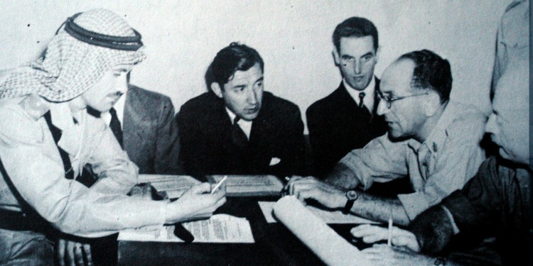 Dated image of six men talking over documents while sitting at a table.
