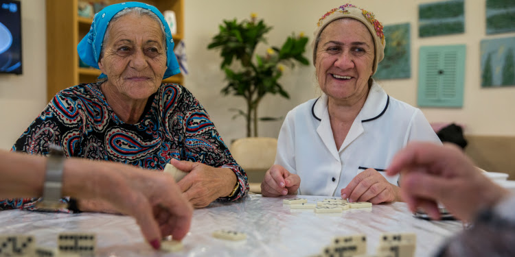 Two elderly women sitting at a table playing dominoes with two other people (not pictured).