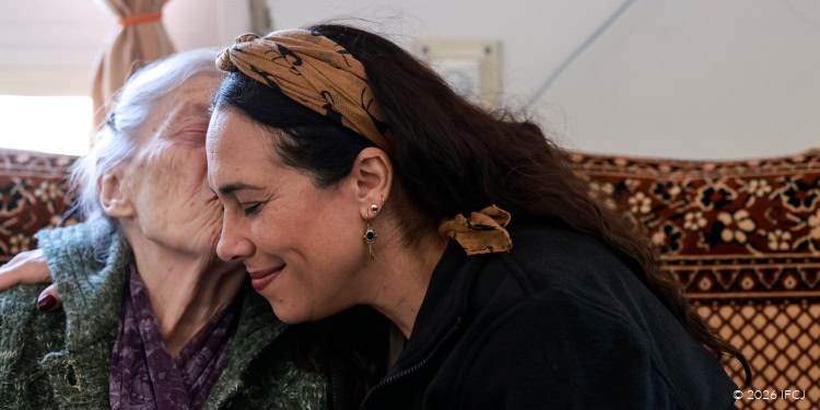 Yael Visiting Bronia Fabrikant - Iran attacks / Operation Roaring Lion (Epic Fury) - March 8, 2026 Yael Eckstein, black branded logo jacket, brown headband - elderly woman, gray knit cardigan, purple scarf - kissing Yael on the cheek - sitting on a couch with a brown and yellow floral design -
