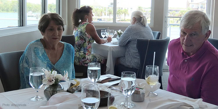 Elderly people enjoying a meal together at a restaurant with large windows and natural light, engaging in conversation.