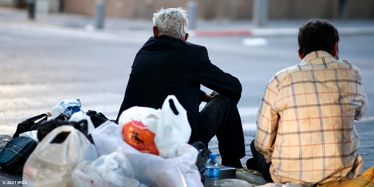Elderly man in street sitting next to young man with several plastic bags behind them.