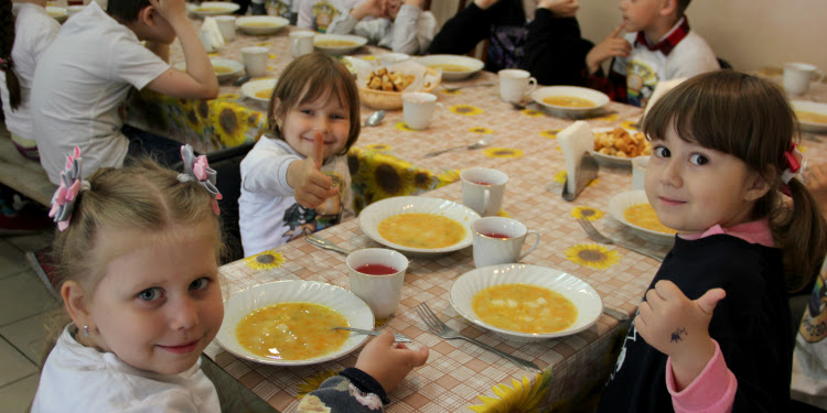 Three young girls eating soup while giving a thumbs up to the camera.
