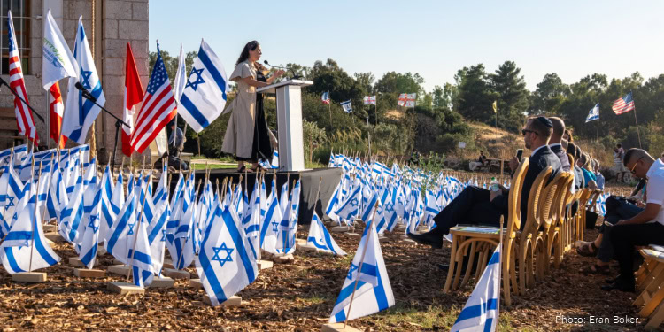 Woman speaking at outdoor event with Israeli flags and audience members seated on chairs, demonstrating support for Israel.