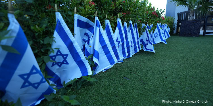 Multiple Israeli flags lined up outdoors on a grassy area with greenery and outdoor furniture in the background.