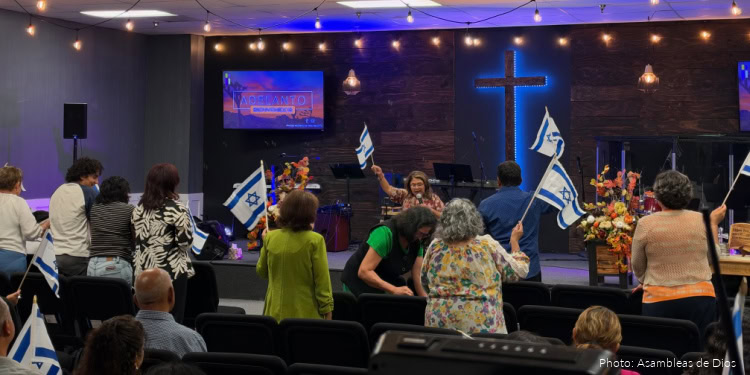 People holding Israeli flags during a fellowship event in a church setting with a cross on the wall and stage lighting.