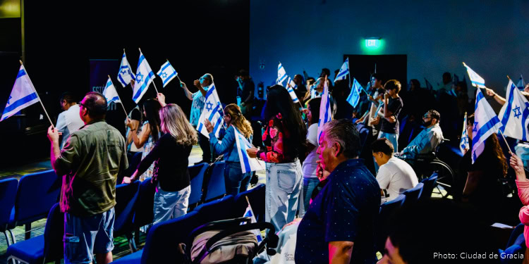 Group of people holding Israeli flags at a fellowship event, with some seated and others standing, in a dark auditorium setting.