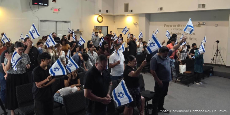 Group of people holding Israeli flags during a fellowship event, showing unity and support for Israel, at an indoor venue with a diverse crowd.