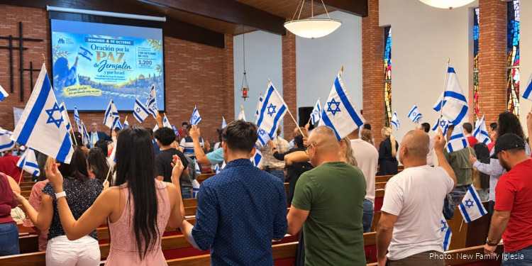Crowd of people holding Israeli flags at a fellowship event, with banners and a large screen in the background.