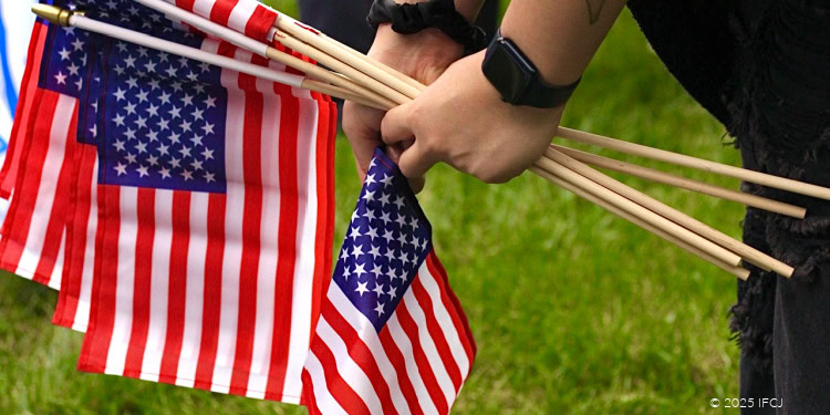 The image depicts an individual holding multiple American flags during an outdoor patriotic event on a green grassy field, emphasizing themes of national pride, unity, and community support.