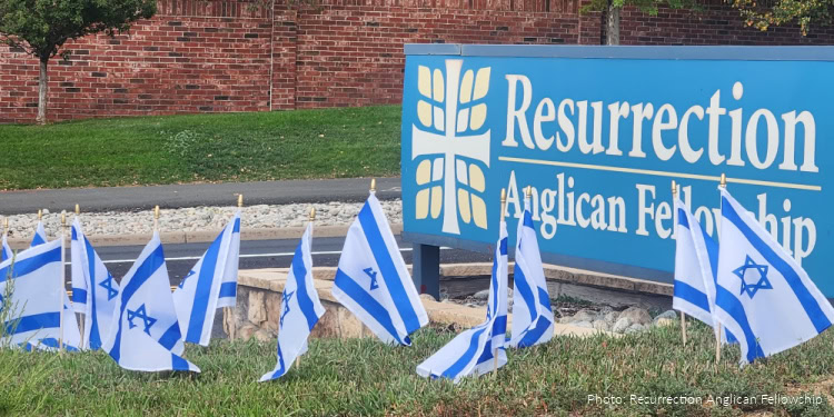 Several Israeli and American flags are arranged outdoors in front of a Resurrection Anglican Fellowship sign, representing international fellowship and religious community. The scene is set on a grassy area with a brick wall and trees in the background, highlighting the celebration of faith and unity among diverse nations.