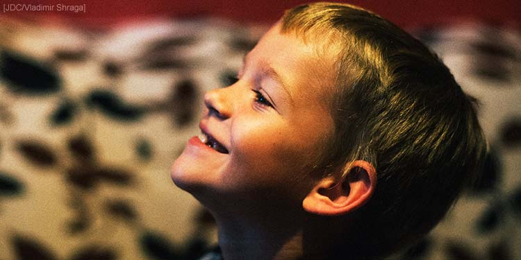 The side profile of a young boy smiling at the air while he stands next to a white and black patterned background.