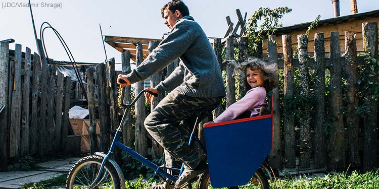 A father riding with a bike that has a blue carriage attached that's holding his daughter.