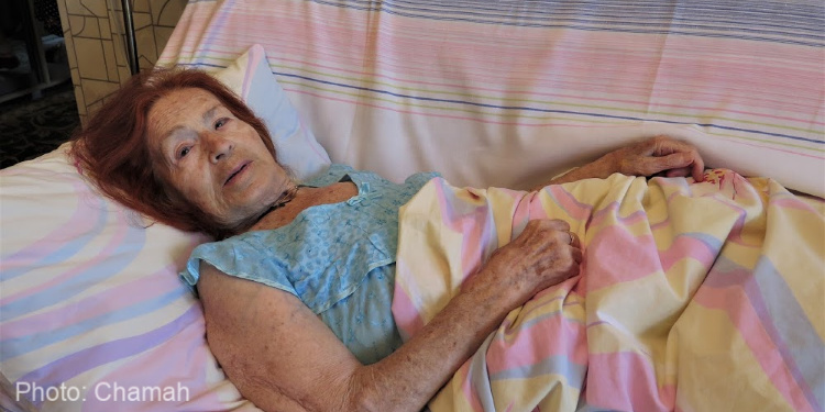 elderly woman lying on bed, blue dress, pink and yellow blanket