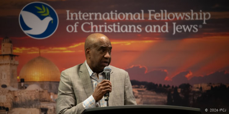 A Black Christian leader speaking into a microphone at an IFCJ event with a Jerusalem backdrop and the International Fellowship of Christians and Jews logo.