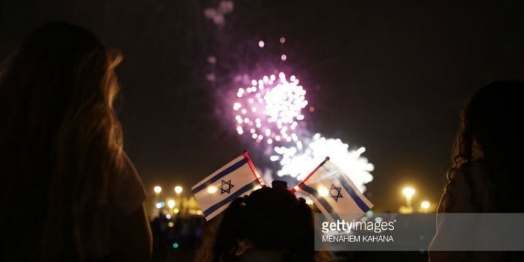 Two young girls holding small Israeli flags in their hands as fireworks shoot off in front of them.