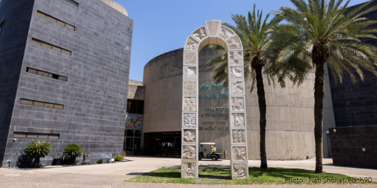 Photograph of a modern building with a stone archway mosaic and palm trees, showcasing an exhibit related to ancient synagogue mosaics.