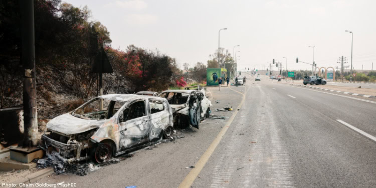 Burned and damaged cars along the roadside in Sderot, with signs of fire damage and debris, under a cloudy sky, illustrating the impact of conflict.