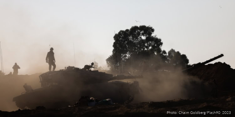 Israeli soldiers and tanks operate in a dusty, conflict zone with smoke and trees in the background, reflecting ongoing military preparedness.