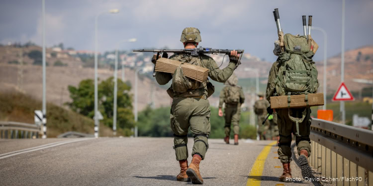 Military personnel walking on a road with backpacks and rifles, in a rural outdoor setting.