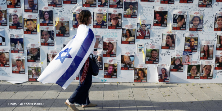 Woman walking with an Israeli flag wrapped around her as she walks by fliers of people who are missing during the Israeli war.
