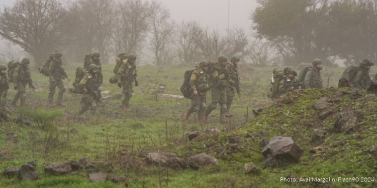 Group of soldiers in military gear walking through a foggy, grassy landscape with rocks and trees in the background.