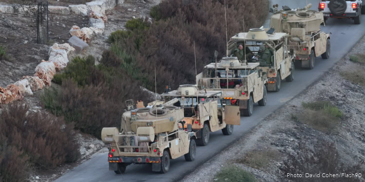 A convoy of military vehicles traveling along a rural road with rocky terrain and sparse vegetation.