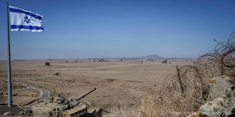 Israeli military tank positioned in a dry, open landscape in southern Lebanon with an Israeli flag flying nearby, under a clear blue sky.