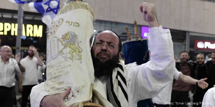 Man celebrating Shemini Atzeret with prayer shawl and Torah scroll in a lively outdoor setting.