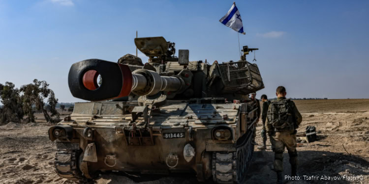 Israeli Defense Forces tank in desert terrain with soldiers nearby, under clear blue sky, during military operation.