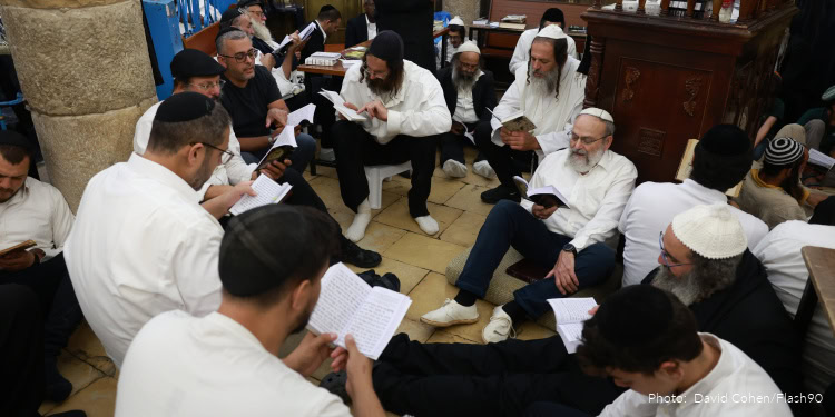 Group of Jewish men praying and reading scriptures during Tisha B’Av observance, sitting on the floor in a synagogue setting.
