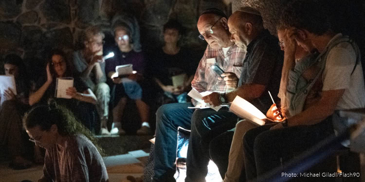 Group of people gathered outdoors at night, lighting candles and reading, commemorating Tisha B’Av, a Jewish day of mourning.
