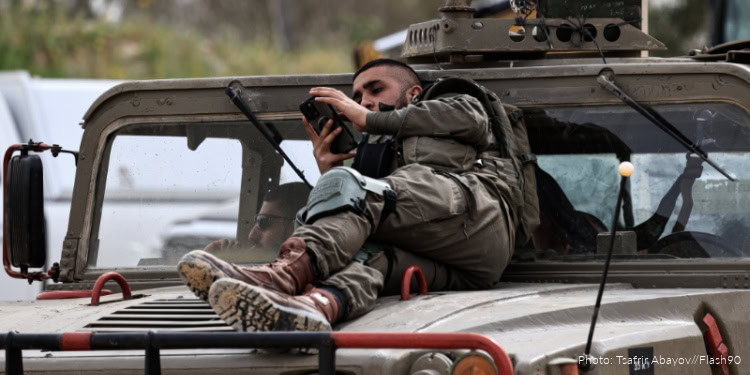 A man resting on top of a military vehicle, using a smartphone, in a conflict zone providing aid and support to refugees.