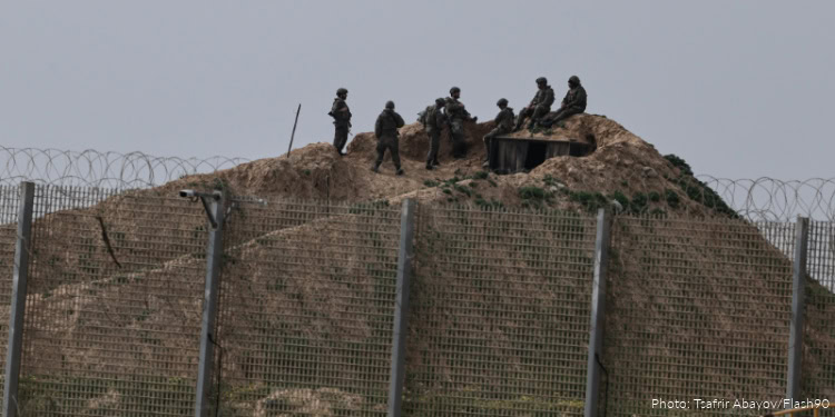 The image shows a group of Israeli soldiers on a mound near the border fence, equipped with military gear and weapons, actively monitoring the surroundings to maintain security and prevent unauthorized crossings.