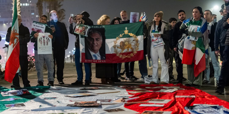 A diverse group of people gather outdoors at night, holding flags, signs, and banners, including an Iranian flag, to show solidarity and support for Iranian issues and human rights.