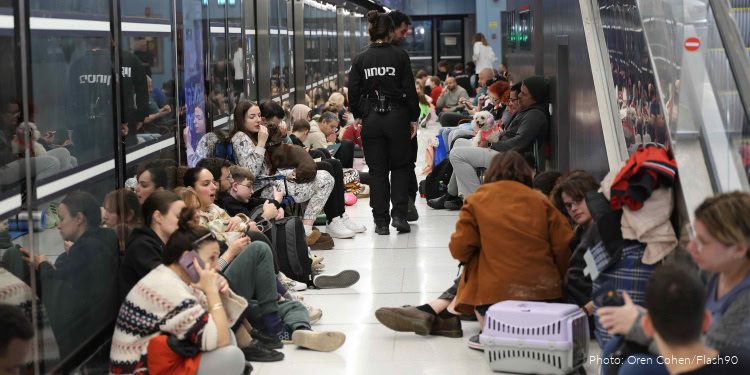 People waiting at an airport terminal, seated along the glass walls, with a security officer walking through the crowd.