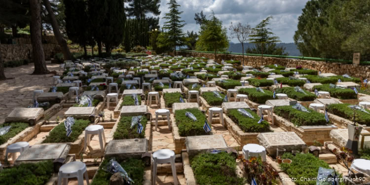Graves at a cemetery with white stools, flowers, and memorial stones, surrounded by trees and a scenic landscape, honoring fallen IDF reservists amid ongoing conflict.