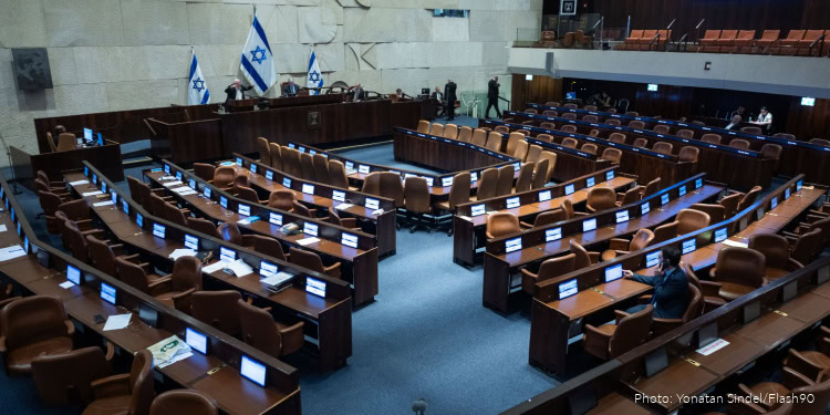 Interior view of the Israeli Knesset assembly hall with empty seats and Israeli flags, symbolizing government discussions and political decision-making.