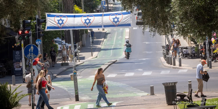 Pedestrians crossing a city street decorated with Israeli flags, with some people walking and others on bicycles, in a lively urban setting.