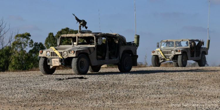 Two military vehicles driving on a gravel road with a clear sky and trees in the background.