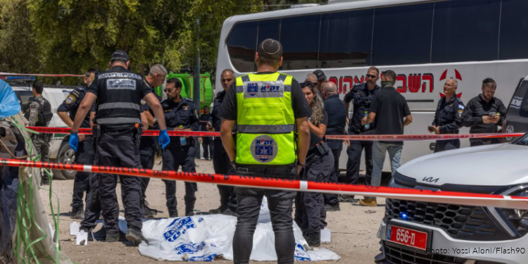 Israeli police officers and emergency responders at a crime scene with a body covered by a white sheet, surrounded by police tape and a bus in the background.