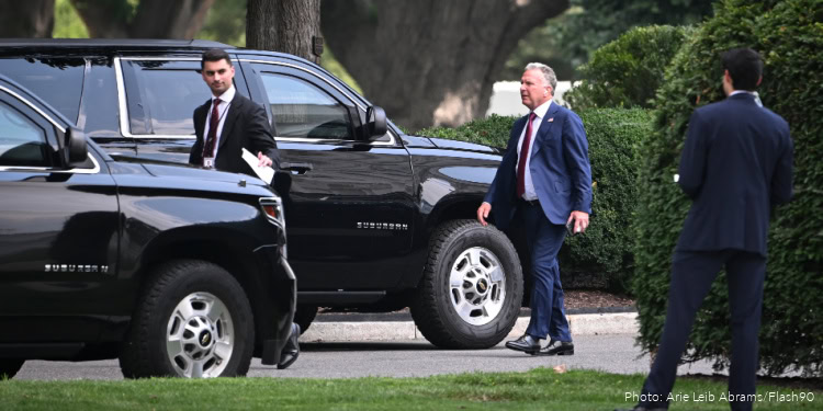 Group of men in suits near black SUVs, possibly diplomats or officials, in an outdoor setting with greenery and trees in the background.