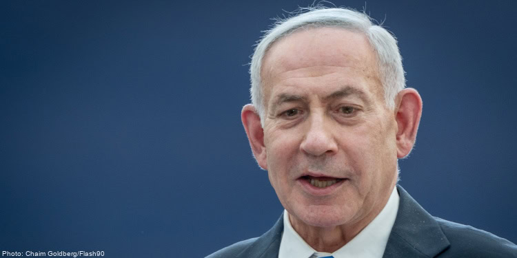 Benjamin Netanyahu, Israeli Prime Minister, speaking at a public event with a blue background, wearing a dark suit and white shirt.