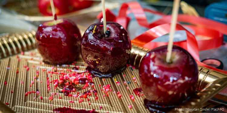 Three shiny candied apples with sticks on a decorative gold tray, surrounded by colorful ribbons and sprinkles, perfect for a festive occasion.